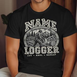 以下が含まれることがあります： 黒いTシャツで、チェーンソーを使って作業する木こり、トラック、丸太が描かれた森のグラフィックプリントが施されています。NAME LOGGER CUT HAUL REPEATという文字が、白とグレーの太いアーチ状のフォントでデザインされています。