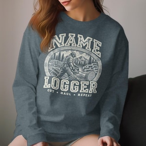 May include: A heathered dark blue long-sleeve sweatshirt featuring a graphic print of a logger using a chainsaw on a log. The text reads NAME LOGGER CUT HAUL REPEAT in a distressed white font, framed by a circular border with a forest background.