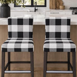 May include: Two backless bar stools featuring a black and white buffalo check fabric upholstery on the square seats. The stools have dark brown wooden frames with four straight legs and a horizontal footrest bar connecting the front legs.