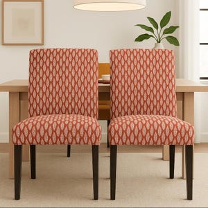 May include: Two armless dining chairs featuring a rust-orange fabric upholstery with a repeating white leaf-like pattern. Each chair has a high, rectangular backrest, a cushioned seat, and four straight, dark brown wooden legs.