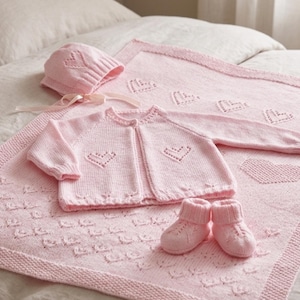 May include: A pink baby blanket, hat, sweater, and booties set. The blanket and sweater feature heart designs. The set is made of a soft, knitted material and is displayed on a bed with a neutral-colored comforter.