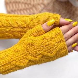 May include: A pair of mustard yellow fingerless gloves. The gloves feature a cable knit pattern on the back and a ribbed cuff. The nails are painted a matching yellow colour.
