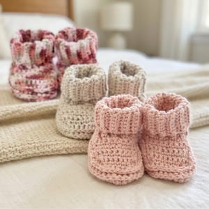 Op de afbeelding: Vier paar handgemaakte gehaakte babyslofjes in verschillende kleuren. Twee paar zijn een mix van roze en wit, één paar is beige en het laatste paar is zachtroze. De slofjes zijn gerangschikt op een wit bed met een crèmekleurige deken.