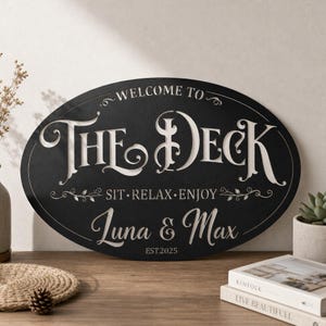 Könnte beinhalten: Ein ovales schwarzes Metallschild mit weißen ausgeschnittenen Schriftzügen, die WELCOME TO THE DECK, SIT RELAX ENJOY, Luna & Max und EST.2025 lauten. Das Schild hat dekorative Verzierungen und eine klare, kräftige Schriftart.