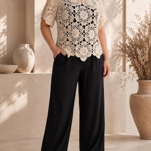 Puede incluir: Una blusa de crochet beige de manga corta con patr&oacute;n floral sobre una camiseta sin mangas negra, combinada con pantalones negros de pierna ancha con bolsillos laterales. La blusa de crochet tiene un cuello y dobladillo festoneados.