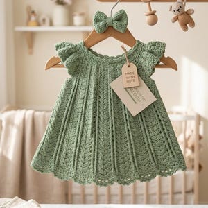 Op de afbeelding: Een saliegroene gehaakte babyjurk met bijpassende strik, hangend aan een houten hanger. De jurk heeft een geschulpte zoom en kapmouwen. Een label leest "Made with Love". Een paar bruinleren babyschoentjes staan eronder.
