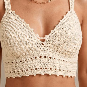 May include: Cream-colored crochet top with a V-neck design. The top features a textured pattern and wide straps. The bottom edge has a scalloped design. The top is made of a soft, natural fiber.