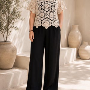 Puede incluir: Una blusa de crochet beige de manga corta con dobladillo festoneado y patrones florales complejos sobre una camiseta sin mangas negra, combinada con pantalones negros de pierna ancha con cintura alta y bolsillos laterales, y sandalias negras.