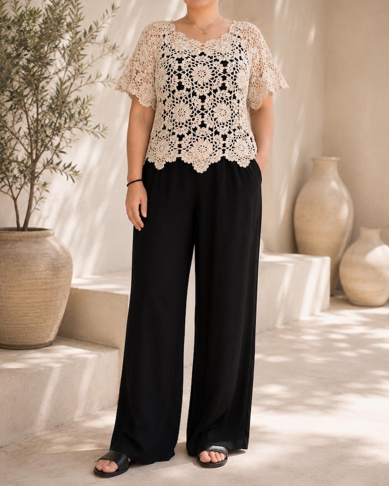 Puede incluir: Una blusa de crochet beige de manga corta con dobladillo festoneado y patrones florales complejos sobre una camiseta sin mangas negra, combinada con pantalones negros de pierna ancha con cintura alta y bolsillos laterales, y sandalias negras.