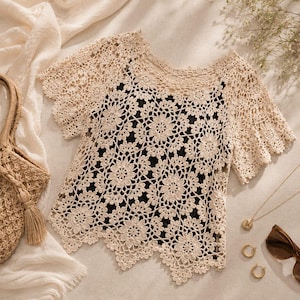 Puede incluir: Un top de ganchillo beige con un patr&oacute;n floral, mangas cortas y un dobladillo festoneado, superpuesto sobre una prenda negra lisa. El top tiene un escote ancho y detalles calados intrincados en toda la tela, creando una apariencia texturizada.
