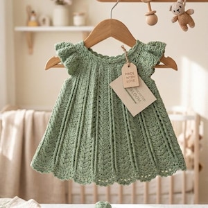 May include: A sage green crochet baby dress hangs on a wooden hanger. The dress has a scalloped neckline and cap sleeves. A tag that says "MADE WITH LOVE" is attached. A pair of tan leather baby shoes and a ball of yarn are in the foreground.