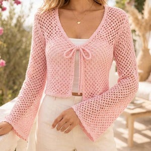 May include: A light pink long-sleeve crochet cardigan featuring an open-knit mesh pattern, a deep V-neckline, and a front tie closure with two thin strings. The garment has flared sleeves and a cropped length, worn over a white top.