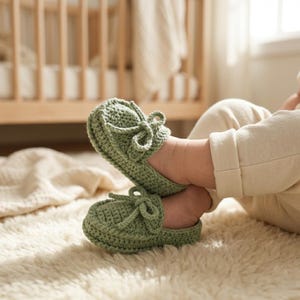 Puede incluir: Un par de mocasines de bebé de ganchillo verde salvia con lazos decorativos. Los zapatos hechos a mano se muestran en los pies de un bebé, con una alfombra suave y texturizada en primer plano y una cuna de madera en el fondo. Los mocasines miden unos 10 cm de largo.