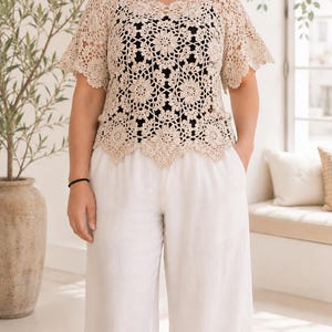 Puede incluir: Un top de ganchillo beige con un patr&oacute;n floral repetitivo, mangas cortas acampanadas y un dobladillo festoneado. La prenda se lleva sobre una capa base negra lisa y se combina con pantalones blancos holgados que tienen bolsillos laterales.