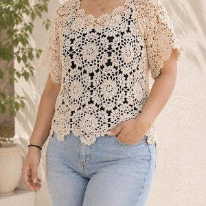 Puede incluir: Una blusa de ganchillo beige con un patr&oacute;n de motivos florales repetitivos, mangas cortas y un dobladillo festoneado, usada sobre una camiseta negra sin mangas y pantalones vaqueros azul claro.