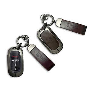 May include: Three car keychains with a dark metallic finish. Two have a leather strap with a debossed logo, and the third has a key fob with buttons. All have a metal ring for attaching keys.
