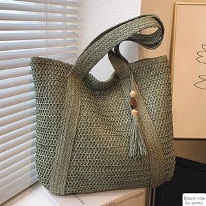 May include: A sage green woven tote bag with braided handles. The bag features a decorative tassel with wooden beads. The bag is made of a textured material and has a rectangular shape. The bag is a stylish accessory.