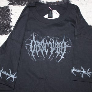 May include: Black long-sleeve shirt with the word "OBSCURA" in a stylized, gothic font across the chest. The sleeves feature a barbed wire design in white. A tag is attached near the collar.