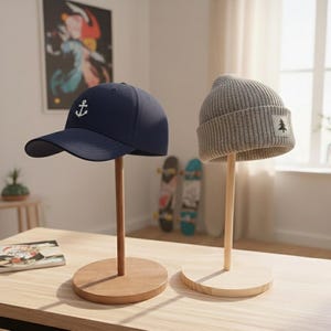 May include: Two hats displayed on wooden stands. A navy blue baseball cap with a white anchor logo and a gray beanie with a small tree patch. The stands have a round base and a vertical wooden post.
