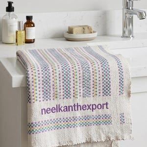 Könnte beinhalten: Ein cremefarbenes Handtuch mit einem gewebten Muster aus vertikalen Streifen in Lila, Grün, Gelb und Blau. Der Text "neelkanthexport" ist in Lila gestickt. Das Handtuch liegt auf einer weißen Arbeitsplatte.