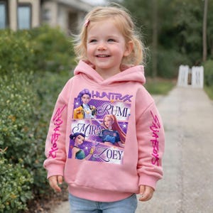 May include: A light pink hooded sweatshirt featuring a graphic of three cartoon characters with the names Rumi, Mira, and Zoey. The word "HUNTRAX" is above the characters. The sleeves have the words "Skija Boys" and "Skija Boys" in pink.
