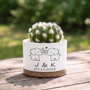 May include: A white ceramic cylindrical planter with a bamboo base containing a green cactus. The planter features a black line drawing of two elephants facing each other with a heart, the letters J & K, and the date 07.14.2026 printed in black.