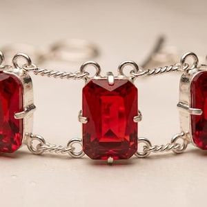 May include: A silver-toned bracelet featuring a series of rectangular, faceted, deep red stones set in prong settings. The stones are connected by double-stranded, twisted silver-toned metal links, creating a structured and repeating geometric pattern.