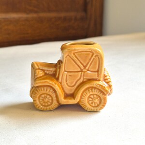 Puede incluir: Pequeña figura de tractor de cerámica de color canela con ruedas y cabina detalladas. El tractor en miniatura tiene una superficie texturizada y está sobre un fondo neutro. La figura es un objeto decorativo.