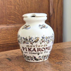 Puede incluir: Jarra de cerámica color crema con tapa, adornada con motivos florales y de pájaros. La jarra presenta el texto "movtarde PIKAROME av vinaigre fin" en burdeos y marrón. Una pieza decorativa para la cocina.