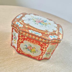 Pode incluir: Uma caixa de lata vermelha e dourada vintage com desenhos florais. A caixa tem formato retangular com bordas arredondadas e tampa articulada. A tampa e as laterais são adornadas com flores coloridas e padrões ornamentados.