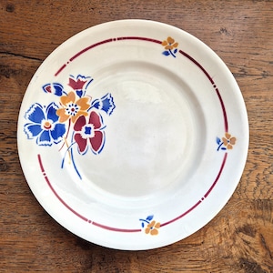 Peut inclure: Assiette en céramique blanche avec un motif floral. L'assiette présente un bord rouge avec de petits tirets et des fleurs stylisées en bleu, orange et rouge. L'assiette est posée sur une surface en bois.