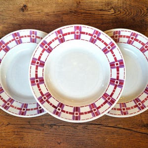 May include: Three white ceramic plates with a decorative red and purple plaid pattern around the rims. The plates are arranged on a wooden surface, showcasing a vintage or retro aesthetic.