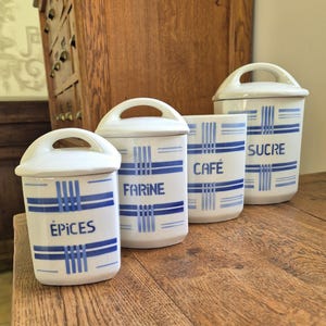 Peut inclure: Quatre pots de cuisine en céramique blanche avec des motifs rayés bleus et des couvercles assortis. Les pots sont étiquetés "ÉPICES", "FARINE", "CAFÉ" et "SUCRE". Ils sont disposés sur une surface en bois.
