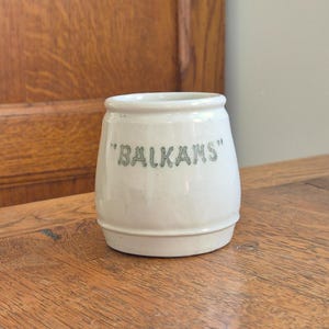 Puede incluir: Un pequeño tarro de cerámica blanquecina con la palabra "BALKANS" en letras verdes. El tarro tiene forma redondeada y está sobre una superficie de madera. El fondo es una puerta de madera.
