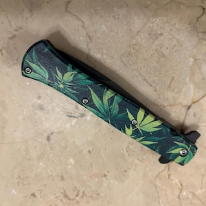 May include: A folding pocket knife with a dark green handle featuring a pattern of green cannabis leaves. The blade is black, and the knife has a black release mechanism and silver rivets.