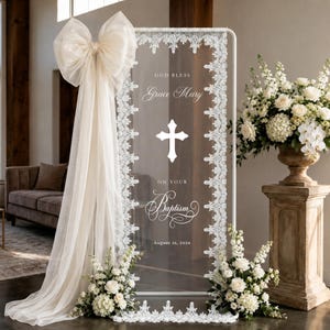 Pode incluir: Uma placa retangular de acrílico transparente com moldura de metal branco, acabamento em renda branca, um grande laço de tecido creme e texto branco GOD BLESS Grace Mary, um ícone de cruz, ON YOUR Baptism e 16 de agosto de 2026.