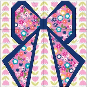 Op de afbeelding: Een stoffen blok met een strik ontwerp. Het midden en de lussen van de strik zijn gevuld met een roze bloemenpatroon, omlijnd in marineblauw. De achtergrond heeft een herhalend patroon van roze tulpen.