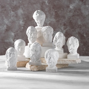 Puede incluir: Colección de bustos de yeso blanco de figuras históricas, como Sócrates y Aristóteles. Los bustos están dispuestos sobre una superficie clara, algunos sobre pequeñas plataformas de piedra. Las figuras tienen rasgos faciales detallados.