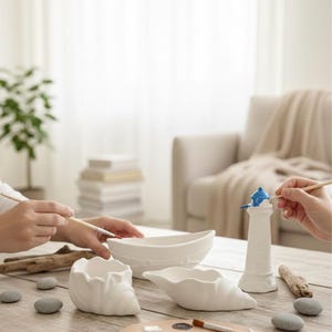 Könnte beinhalten: Eine Bastelszene mit weißen Keramikteilen, darunter eine bootsförmige Schale, ein Leuchtturm und muschelförmige Schalen, die mit Pinseln bemalt werden. Eine Holzpalette mit Farben und kleinen Steinen liegt auf dem Tisch.