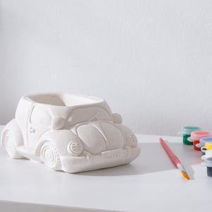 Könnte beinhalten: Ein weißer Keramik-Blumentopf in Form eines Autos mit detailliertem Design, bereit zum Bemalen. Ein Pinsel und kleine Farbtöpfe in verschiedenen Farben liegen neben dem Pflanzgefäß, was auf ein DIY-Bastelprojekt hindeutet. Der Pflanztopf hat ein Nummernschild mit der Aufschrift "9122".