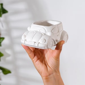Puede incluir: Una maceta blanca con forma de coche. La maceta es una réplica detallada de un coche clásico, con faros, ruedas y una matrícula. La parte superior del coche está abierta, creando un espacio para plantas u otros objetos decorativos.