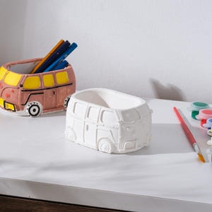 May include: Two ceramic bus-shaped planters, one painted in pink, yellow, and black, holding pencils. The other is unpainted white. Paint pots and a paintbrush are next to the white planter. The planters are on a white surface.