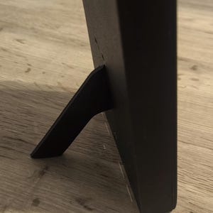 Puede incluir: Un marco de fotos negro con un soporte incorporado. El marco es rectangular y tiene un acabado mate. El soporte está fijado a la parte posterior y permite que el marco se mantenga en posición vertical sobre una superficie plana.