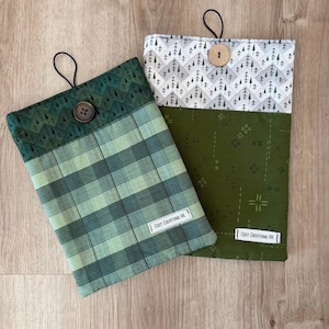 May include: Two fabric book sleeves with button closures. One sleeve features a green plaid pattern, the other has a forest-themed print. Both have a black loop and a label that reads "COZY CREATIONS AK".