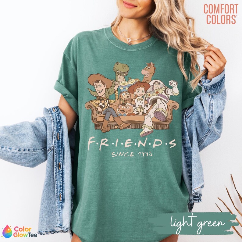 May include: A forest green t-shirt featuring a graphic of Toy Story characters sitting on a couch with the text F.R.I.E.N.D.S SINCE 1995. The shirt has a crew neckline and short sleeves, worn with a light blue denim jacket over the shoulders.
