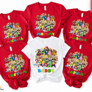 May include: Red and white t-shirts featuring a colorful graphic of characters from Universal Studios. The shirts have names like "Michael", "Mommy", "Marcus", "Custom", "Daddy", and "Sophia" printed on them. The text "Universal Studios" is also visible.