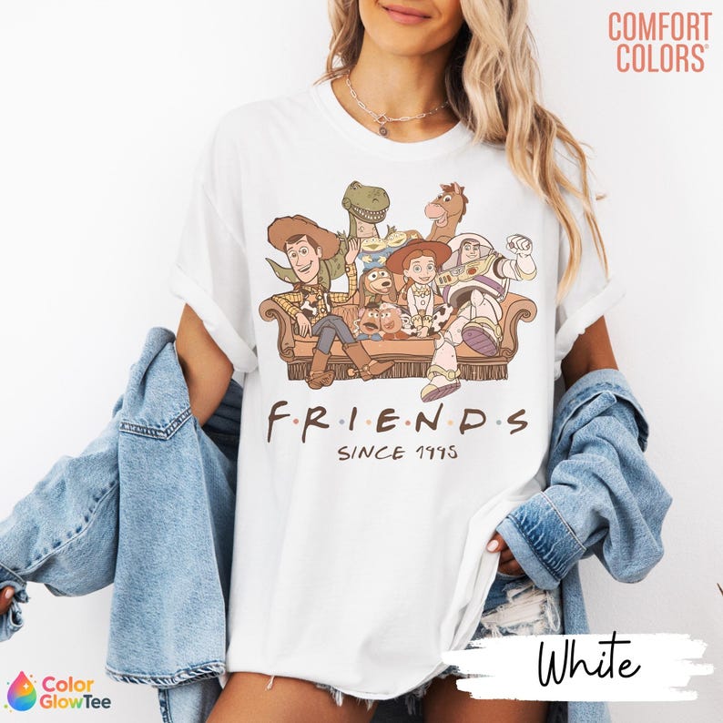 May include: A white short-sleeve t-shirt featuring a graphic print of various animated characters sitting on a couch. Below the characters, the text reads "F R I E N D S" and "SINCE 1995". The top right corner displays the text "COMFORT COLORS".