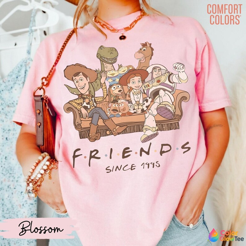 May include: A light pink short-sleeve t-shirt featuring a graphic of Toy Story characters sitting on a couch with the text "FRIENDS SINCE 1995" printed below. The shirt has a crew neckline and is made of a soft cotton fabric with a vintage wash.