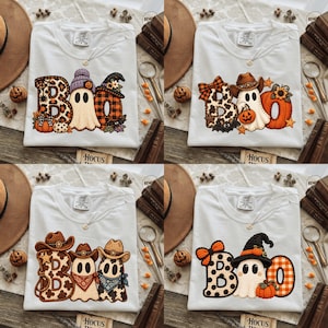 May include: Four white t-shirts featuring graphic prints of the word BOO. The designs incorporate ghosts, pumpkins, cowboy hats, and various patterns like leopard print, plaid, and polka dots in orange, black, brown, and purple color schemes.