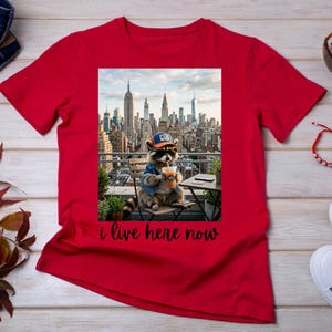 Può includere: Una t-shirt rosso brillante con una stampa grafica di un procione che indossa una giacca di jeans blu, occhiali da sole e un cappellino da baseball NYC, mentre tiene un caffè su un balcone con vista sullo skyline con il testo i live here now.